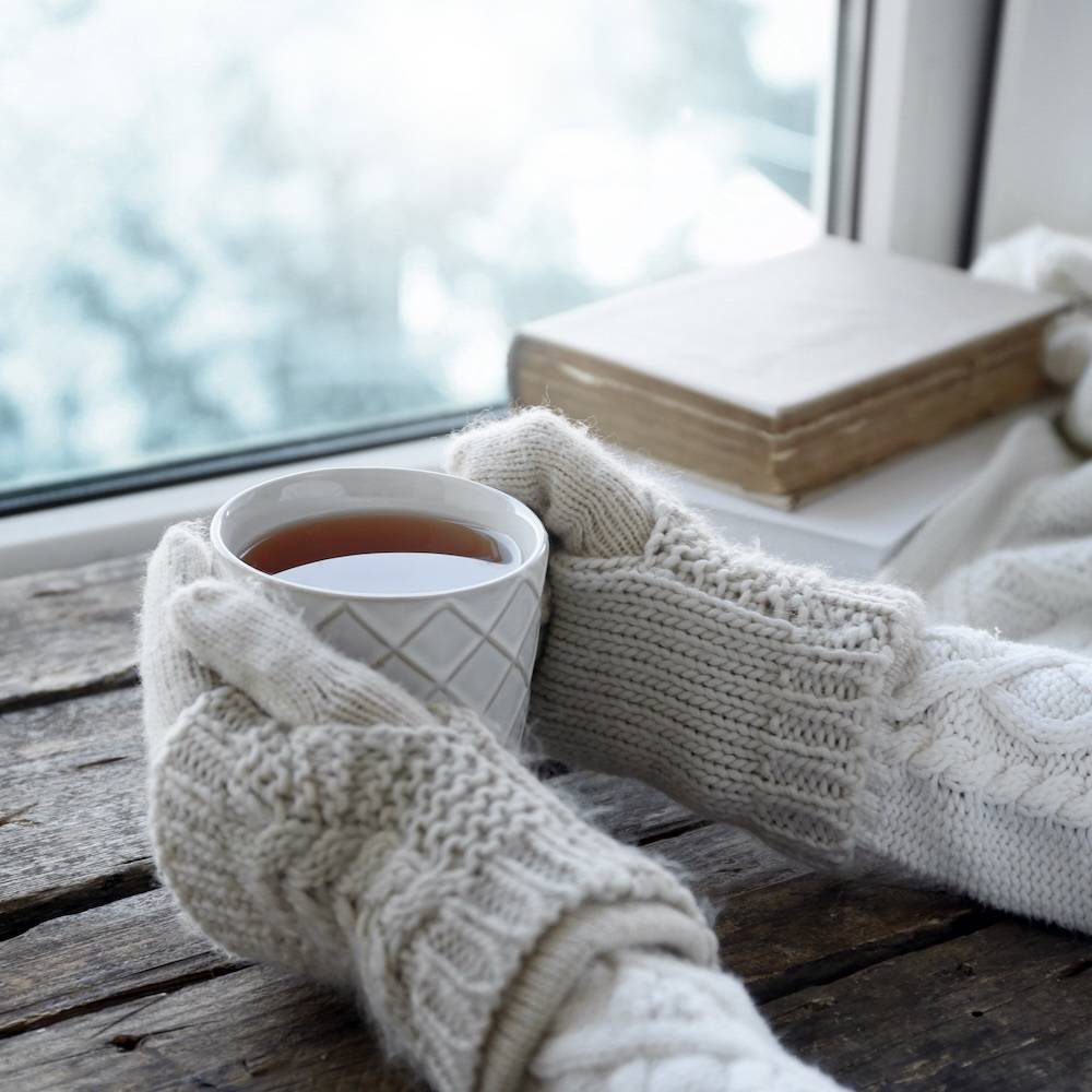 窓際で手袋をしながら紅茶を飲む女性と本