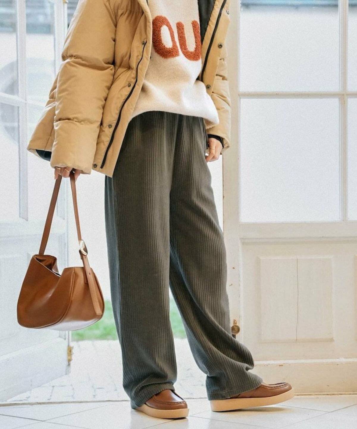 ロゴニットに白パンツを合わせてブラウンのコートを持った女性