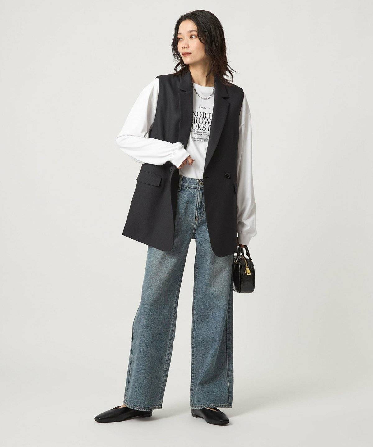 黒トップスにベージュのベストを重ねてデニムを穿いた女性