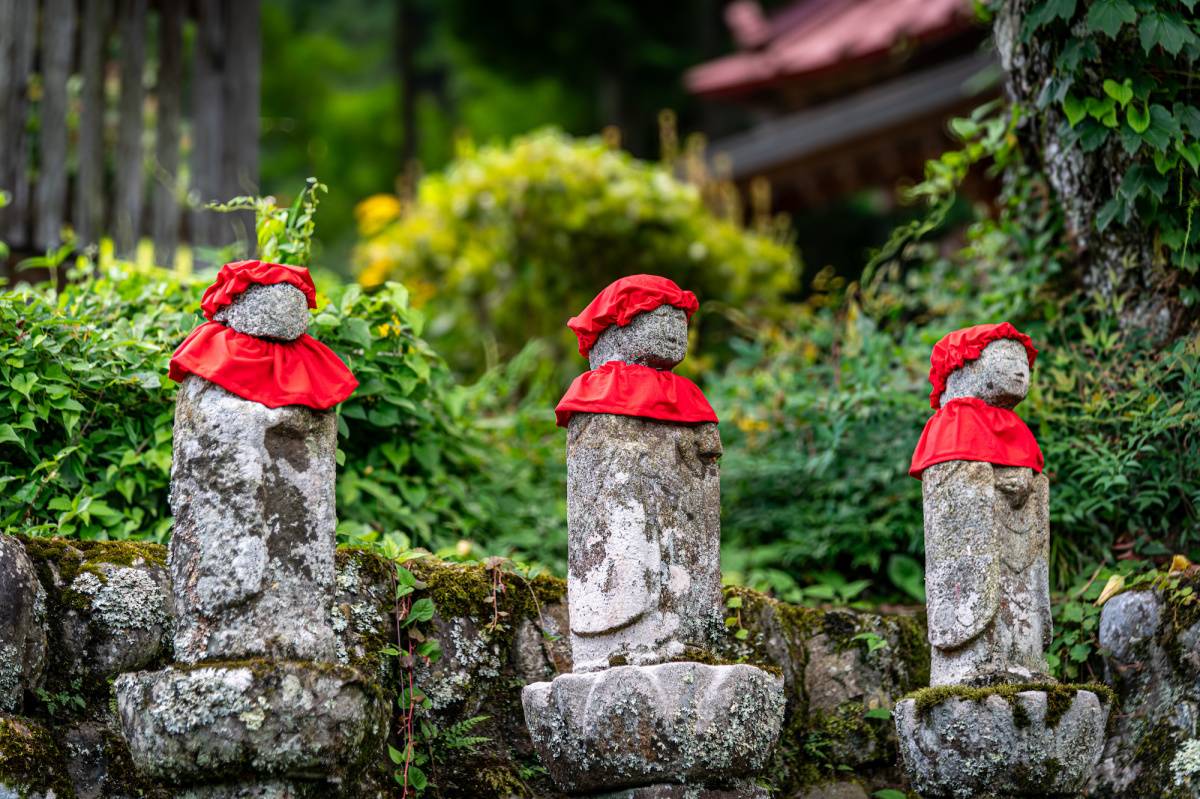 赤い前掛けと帽子をつけた、三体のお地蔵さんが並ぶ田舎道の風景