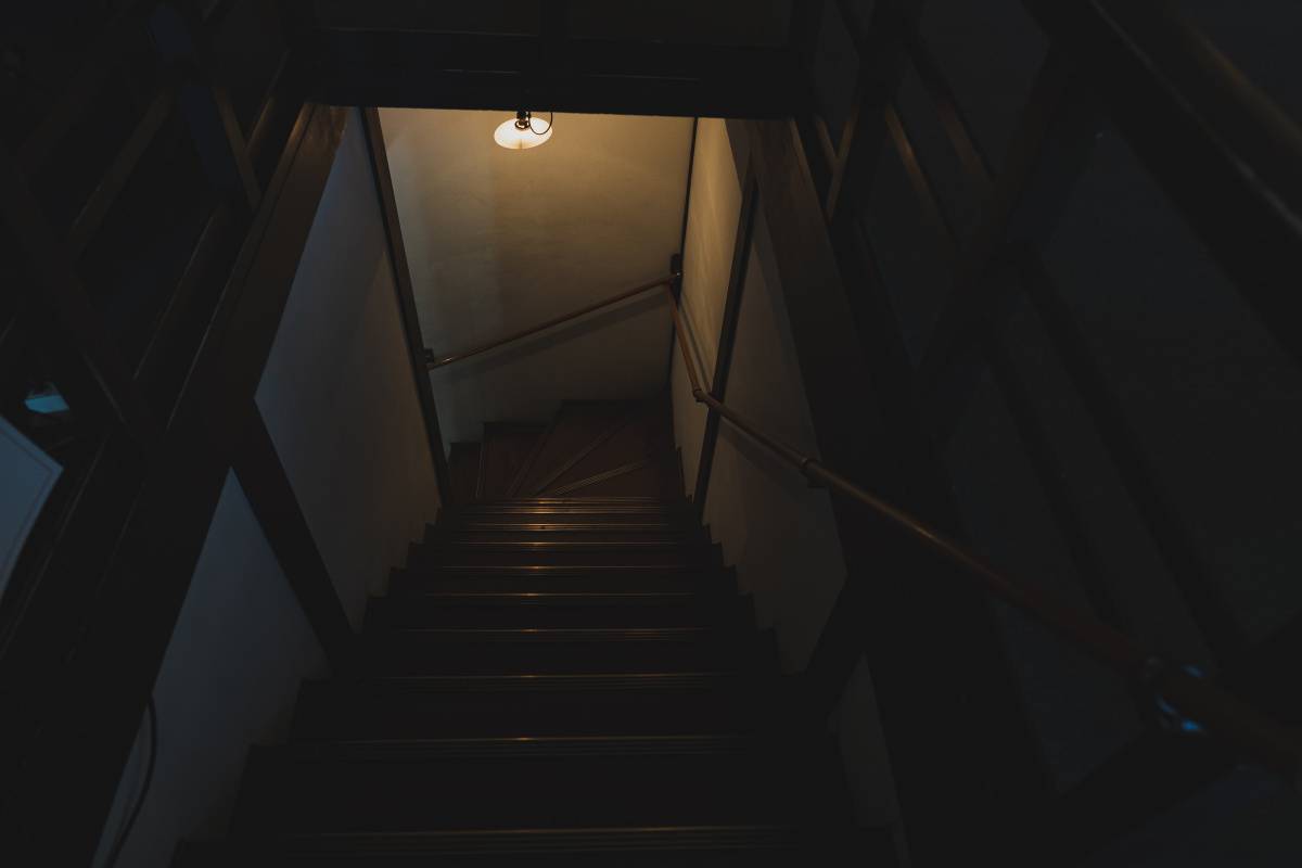 薄暗い古い木造住宅の階段。天井の照明が一つだけ灯り、奥へ階段が続いている。