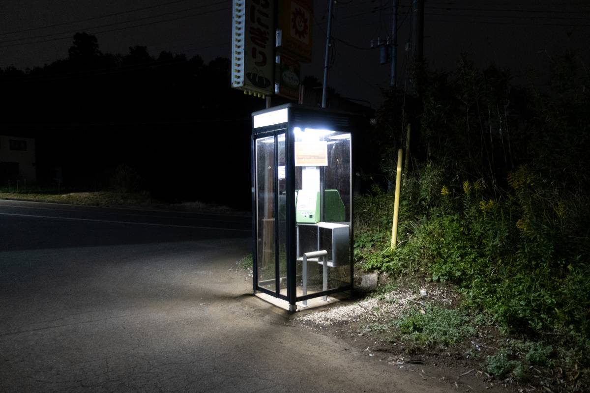 夜の道路脇に設置された公衆電話ボックス。周囲は暗く、電話ボックスの照明だけが明るく灯っている。