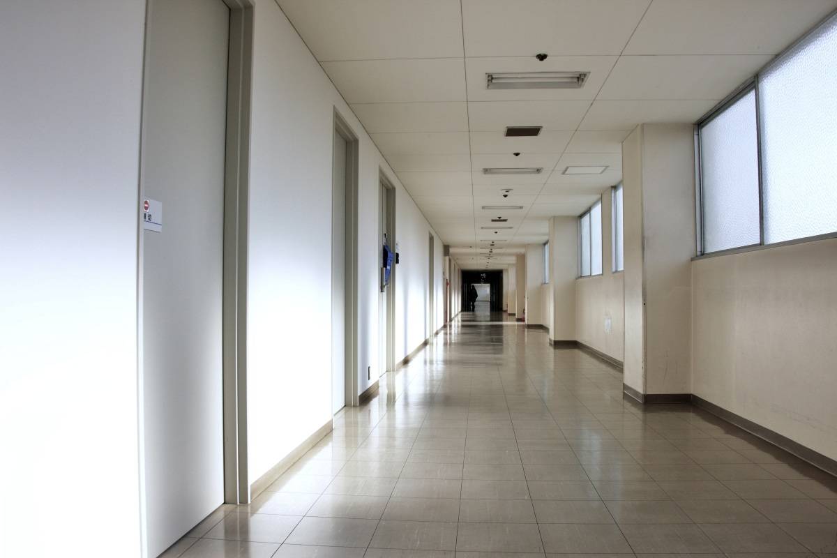 白い壁と窓が続く建物の長い廊下。廊下の奥には小さな人影が見え、突き当たりは薄暗くなっている。
