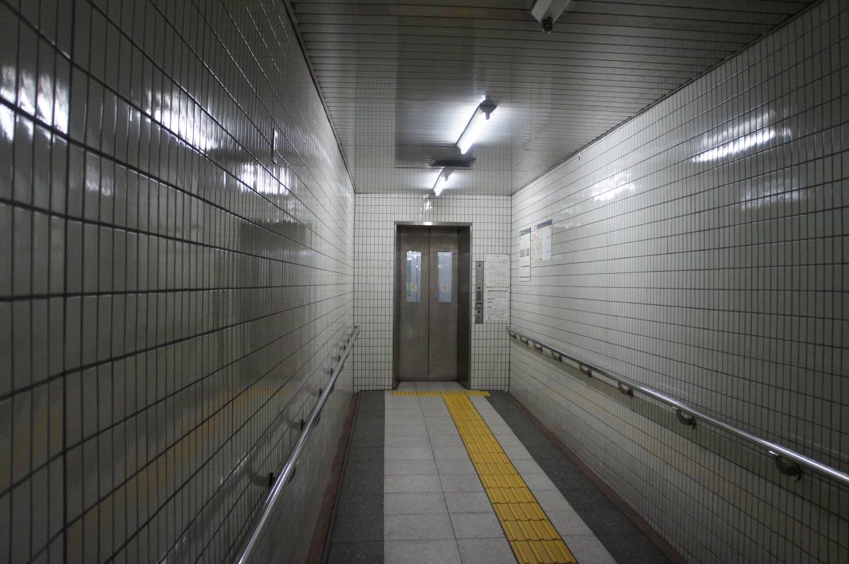 白いタイル張りの地下通路の奥にあるエレベーター。床の黄色い点字ブロックが扉の前まで続いている。