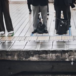  駅の向かい側のホームに並ぶ乗客たち。足元にはバッグが置かれ、ホームの端には点字ブロックが続いている。 
