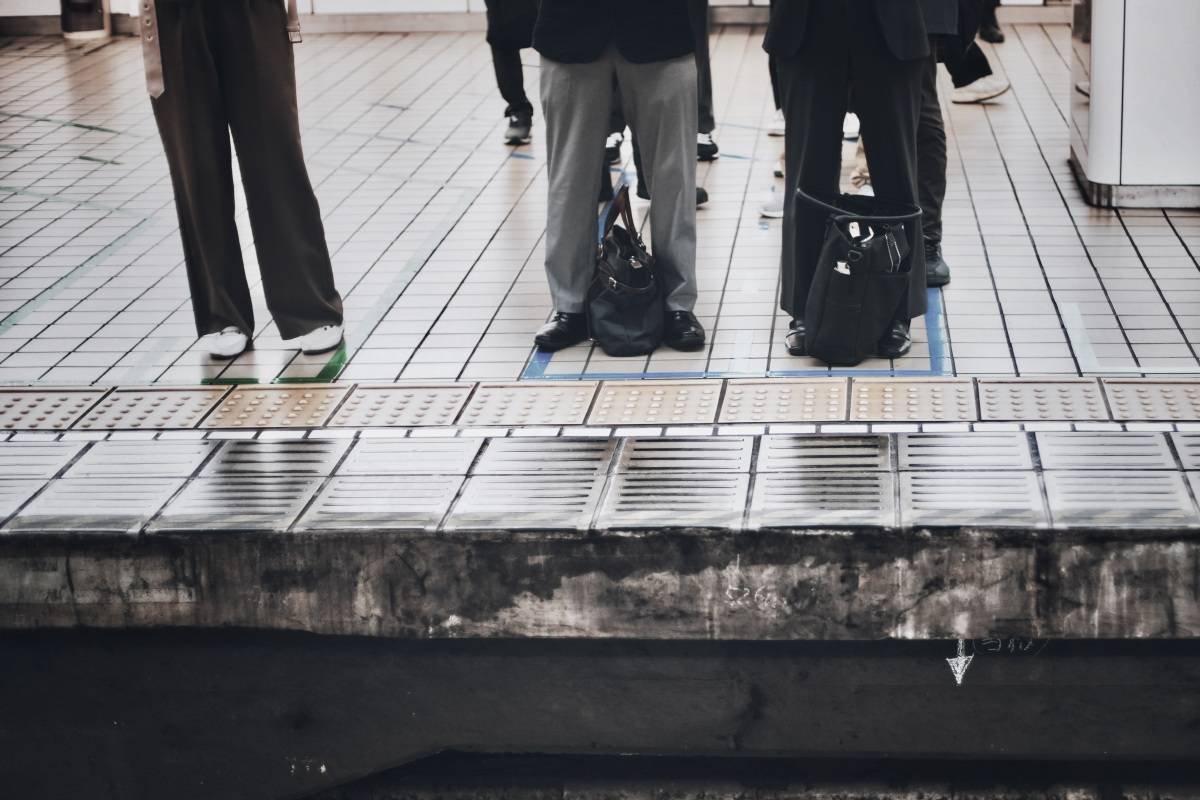 駅の向かい側のホームに並ぶ乗客たち。足元にはバッグが置かれ、ホームの端には点字ブロックが続いている。
