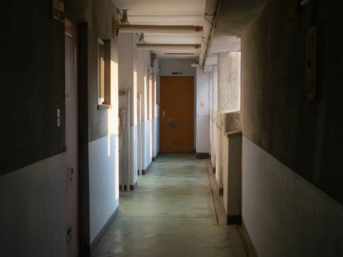 古い集合住宅の廊下。両側に玄関や配管が並び、突き当たりに茶色いドアがある。